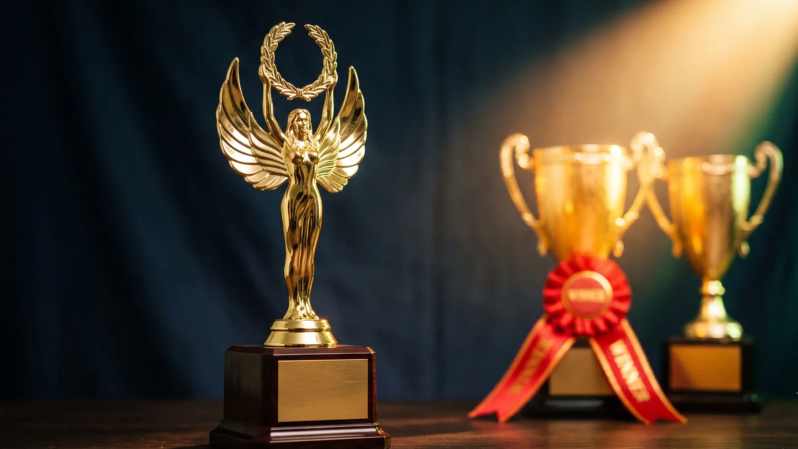 A gold winged-victory trophy in sharp focus in the foreground while a warm spotlight and a red winner's ribbon fall on two other trophies behind it